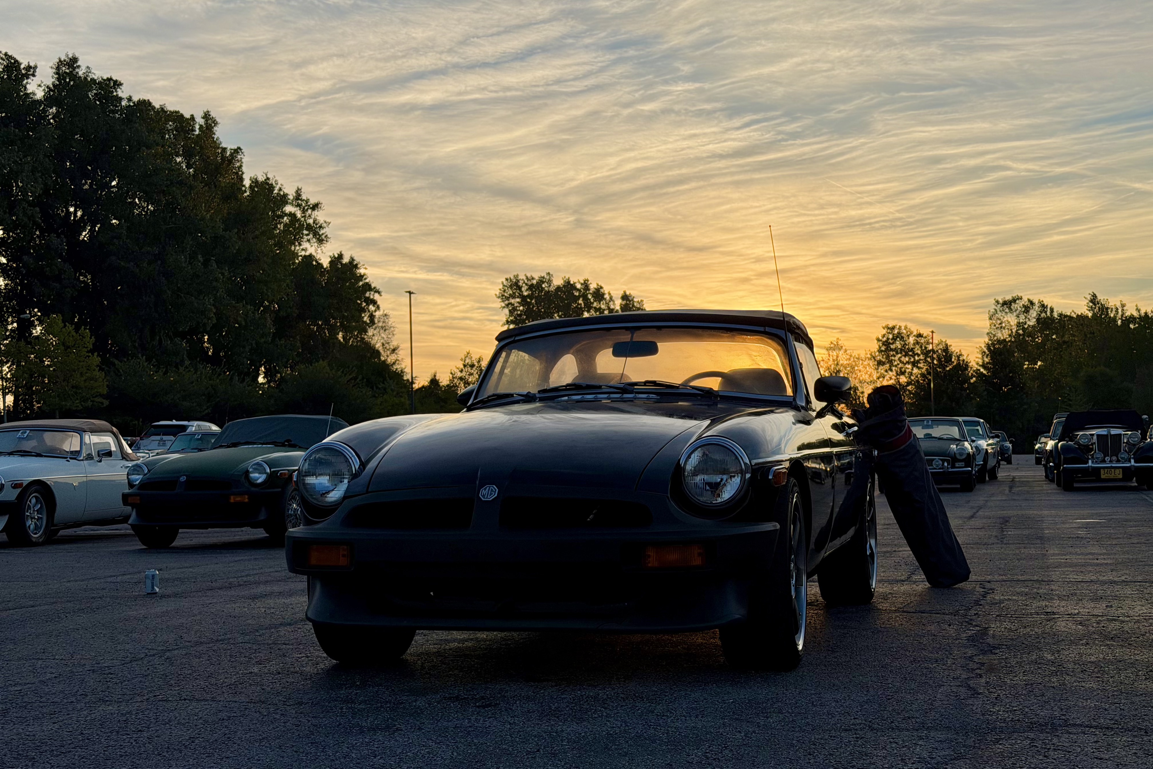 Photograph of a black 1976 MGB from the front with a sunrise behind in a parking lot full of MGs.