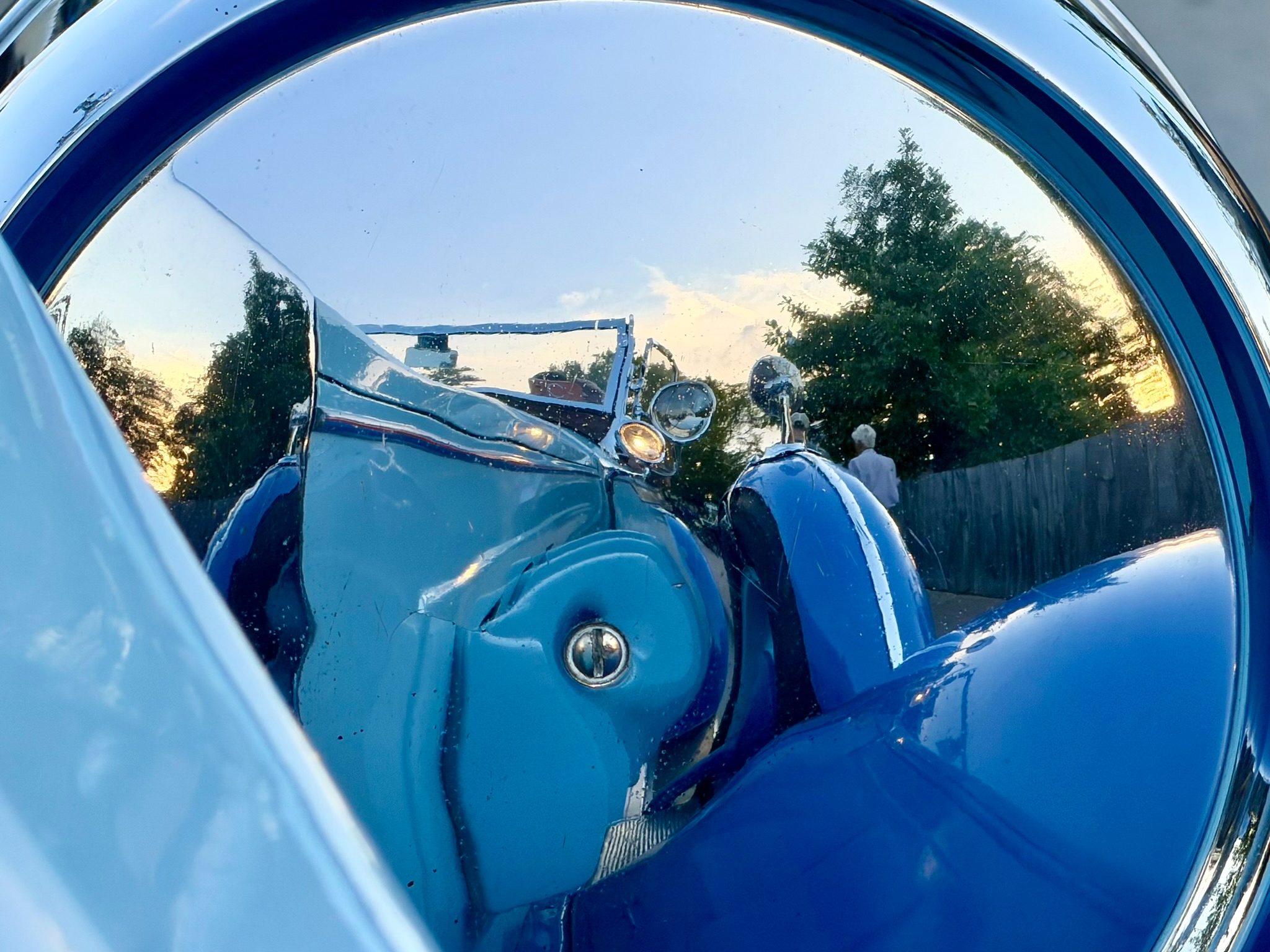 View of the passenger side of a periwinkle blue 1928 Packard 443 Custom Dual-Cowl Phaeton as seen reflected and distorted in the chrome rear of its passenger-side headlamp.