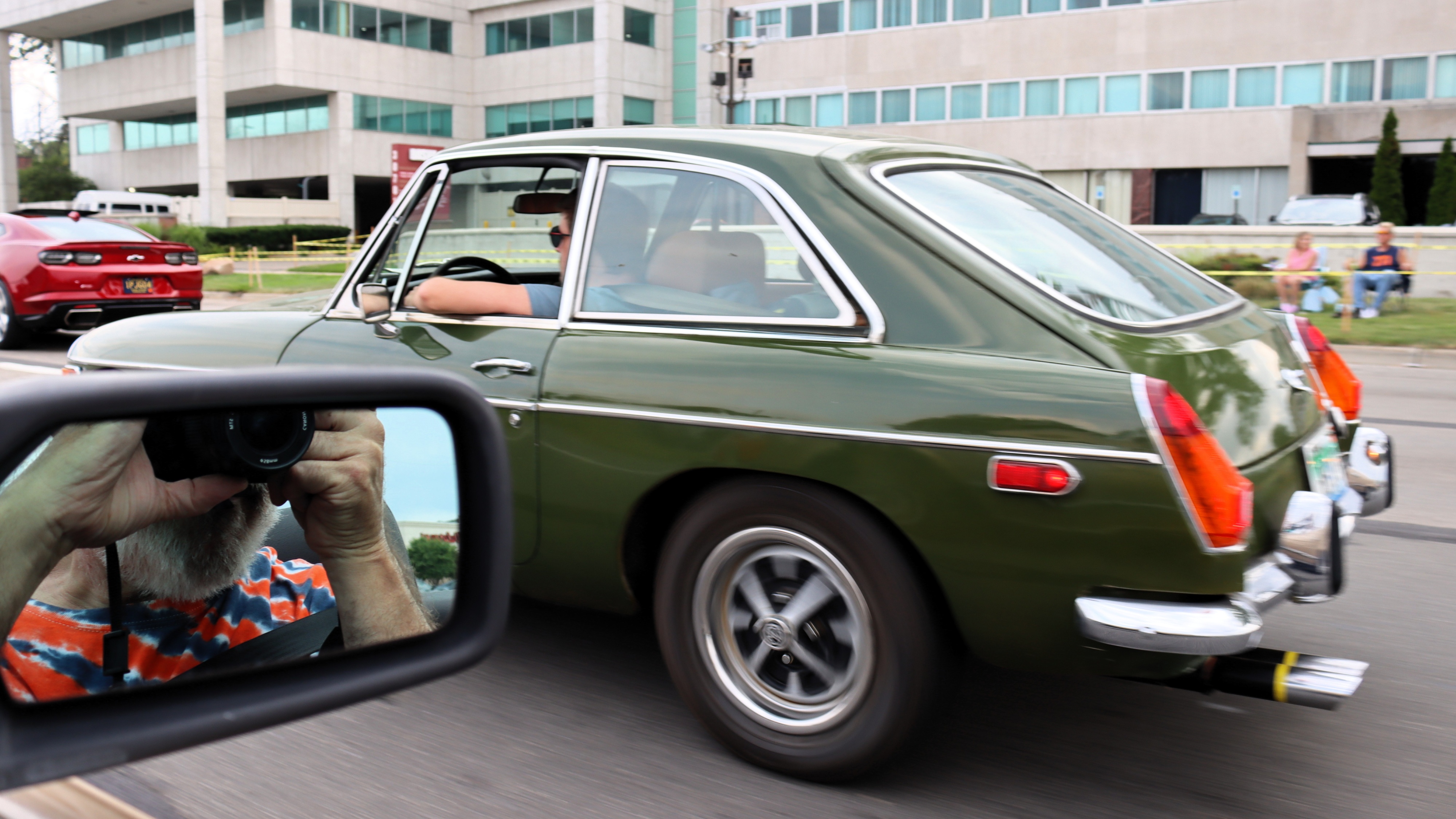 Photograph from the side of an early 1970s tundra green MGB GT coupe with the photographer caught in the rear-view mirror of his own car in the lower right of the image, completely blocking the front left of the MGB GT.