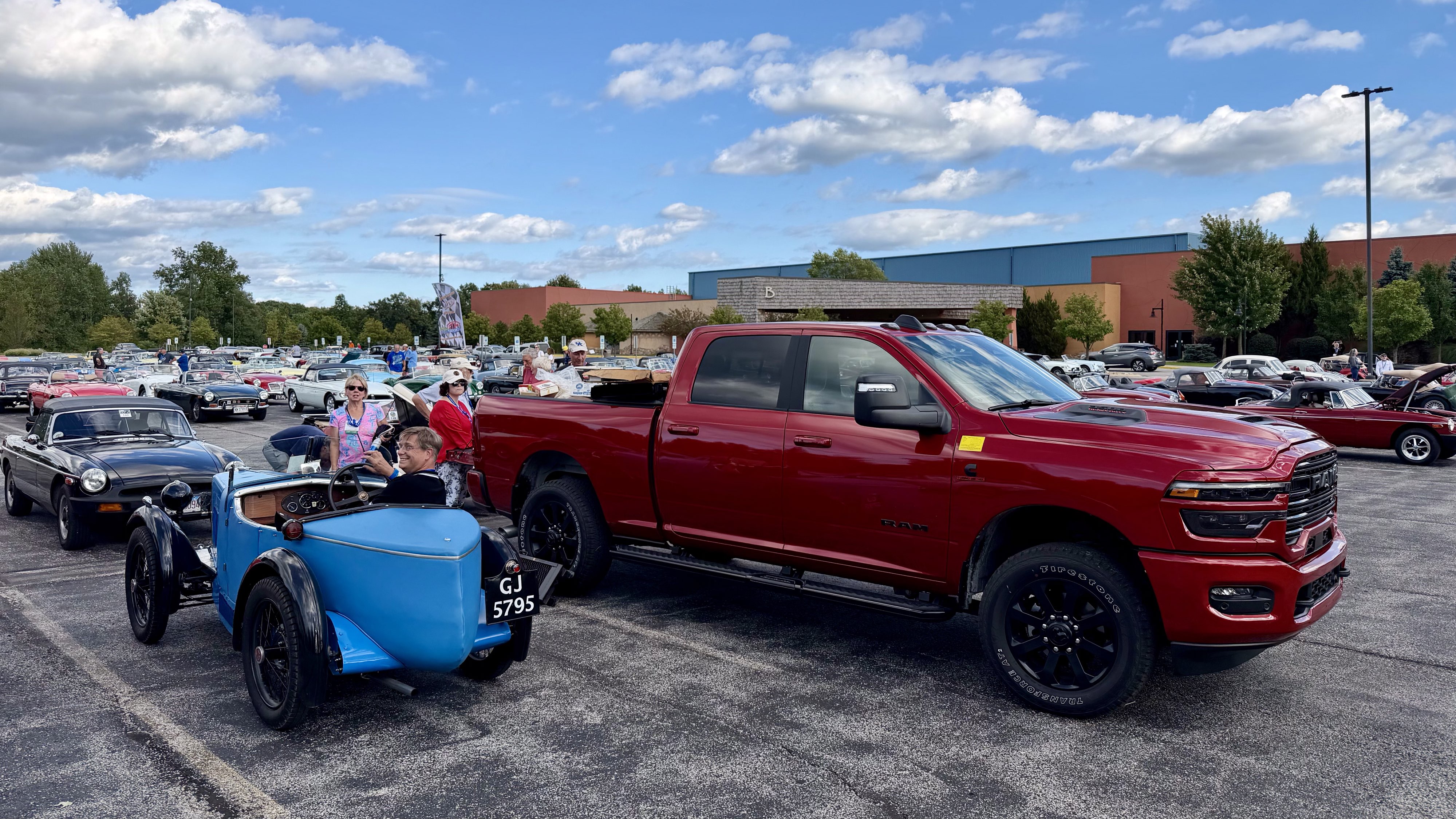 Photograph of a small blue 1930 MG M-Type next to a large red Ram pickup in a parking lot full of MGs at MG International 2026 in Sandusky, Ohio.
