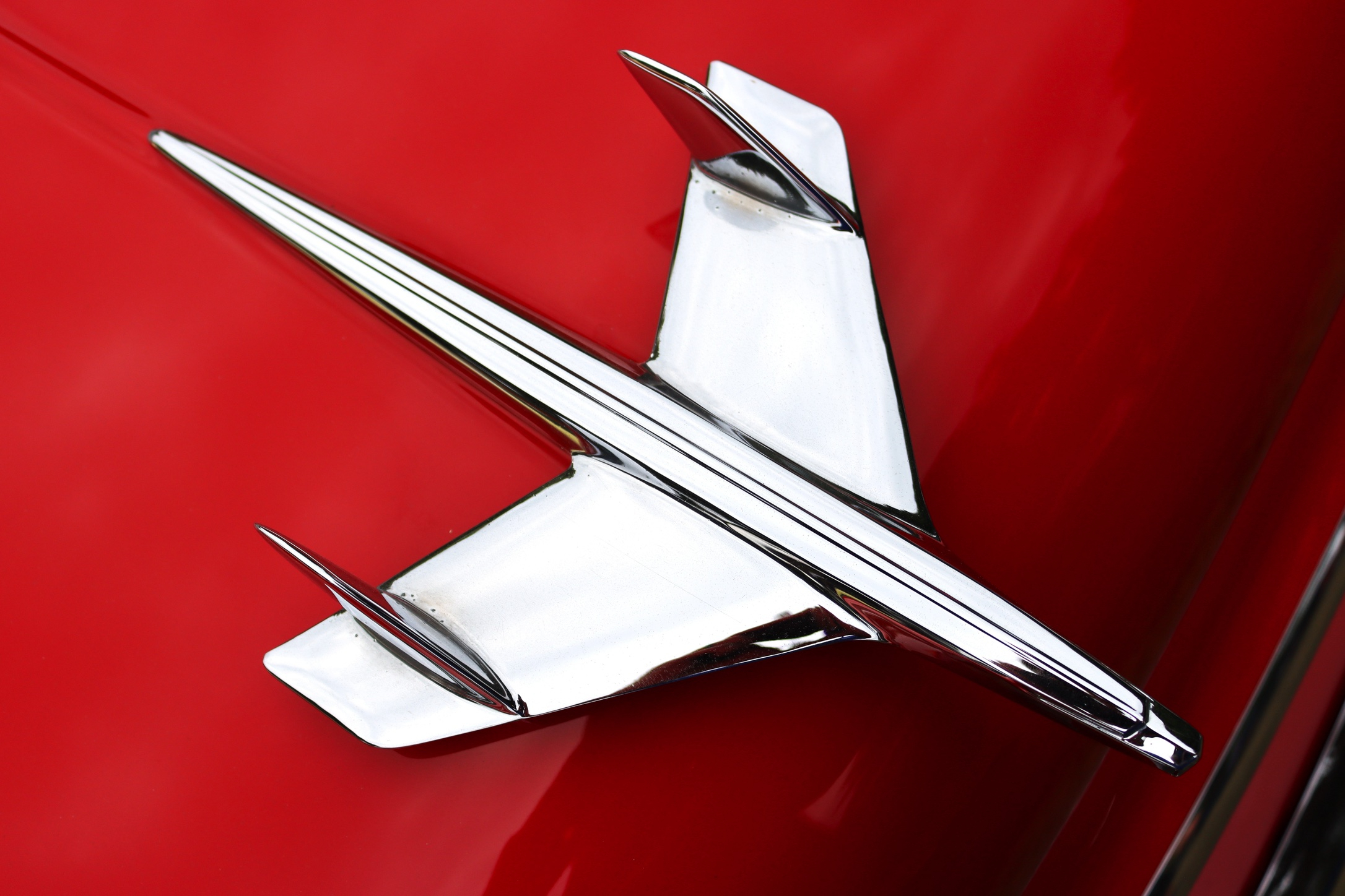 Photograph from above of an early 1950s Chevy airplane-style chrome hood ornament on a red Chevrolet automobile.