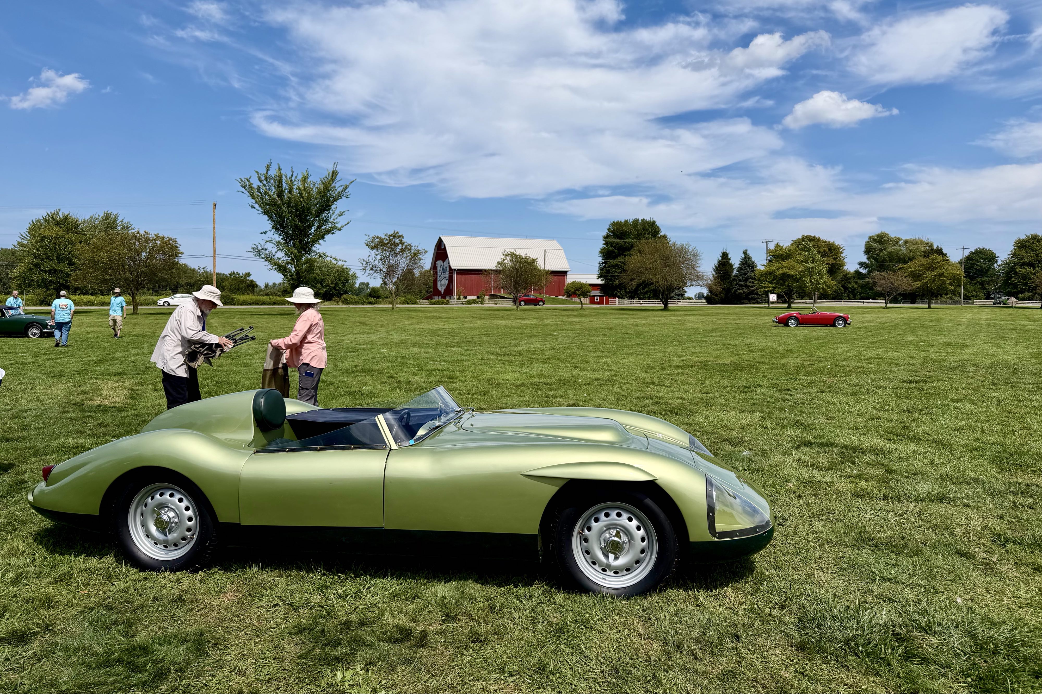 Photograph of a sleek green open top racing car from the late 1950s on a grass field with a barn behind as the car show clears after the car show at MG International 2026 in Sandusky Ohio.