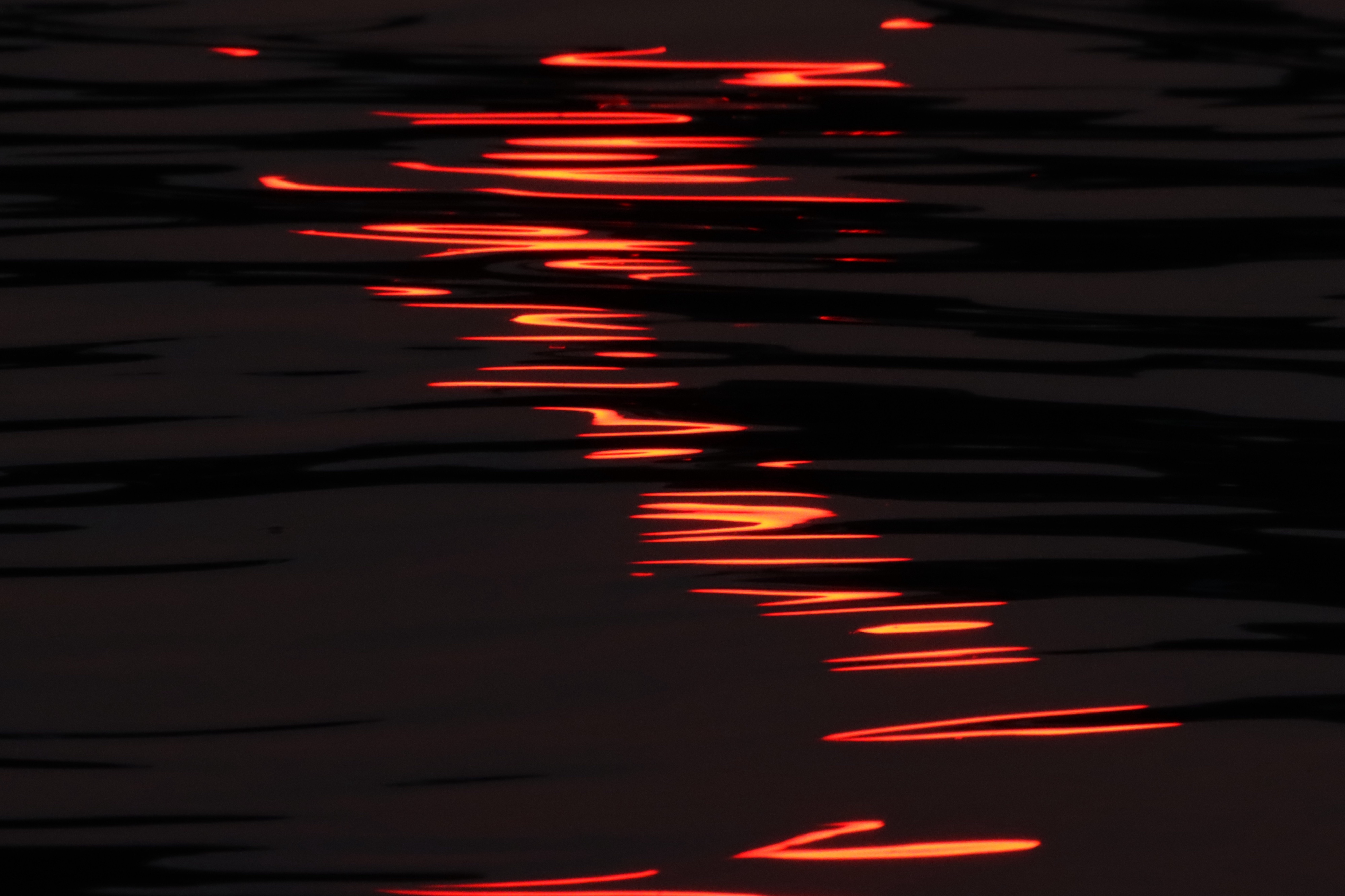 Photograph of orange sunlight reflected on the ripples of dark water, forming an elegant swoop of shapes going down the middle of the image.￼