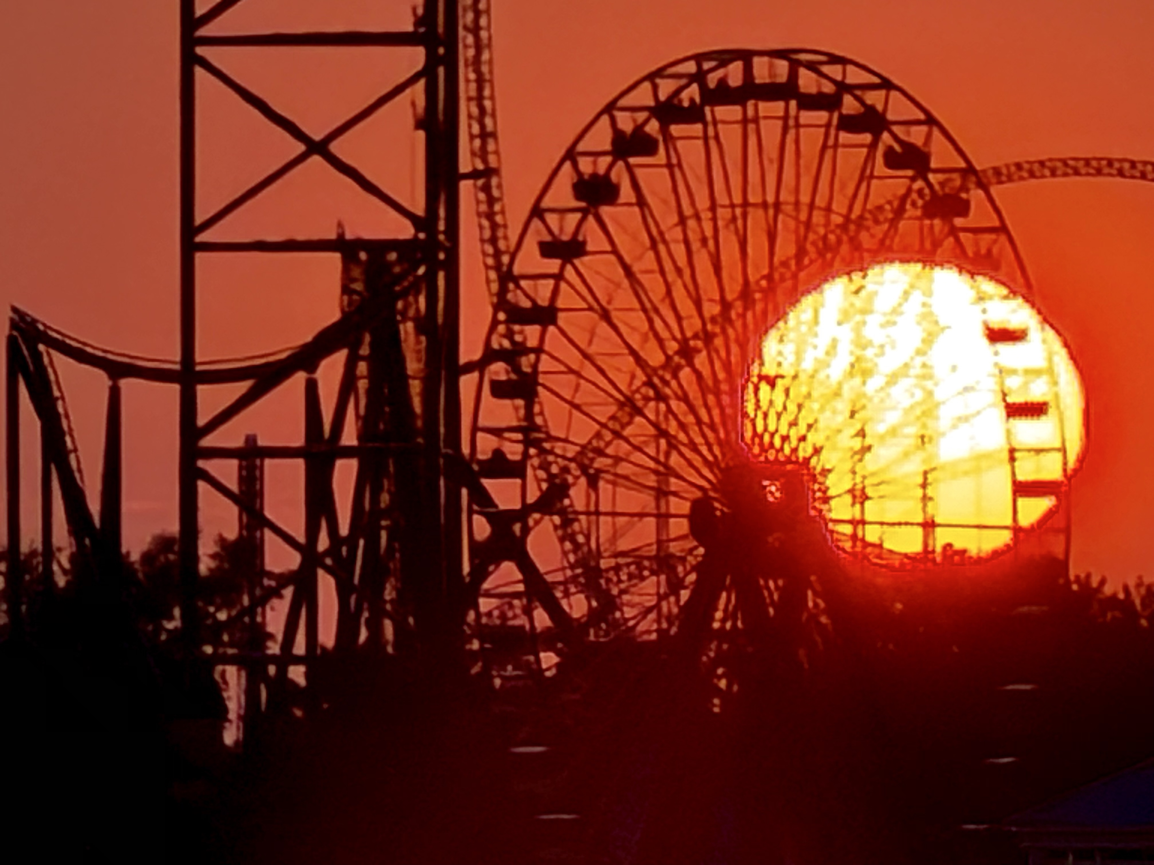 Sunset photograph of a large yellow Sun in an orange sky with a large Ferris wheel and other rides in silhouette.