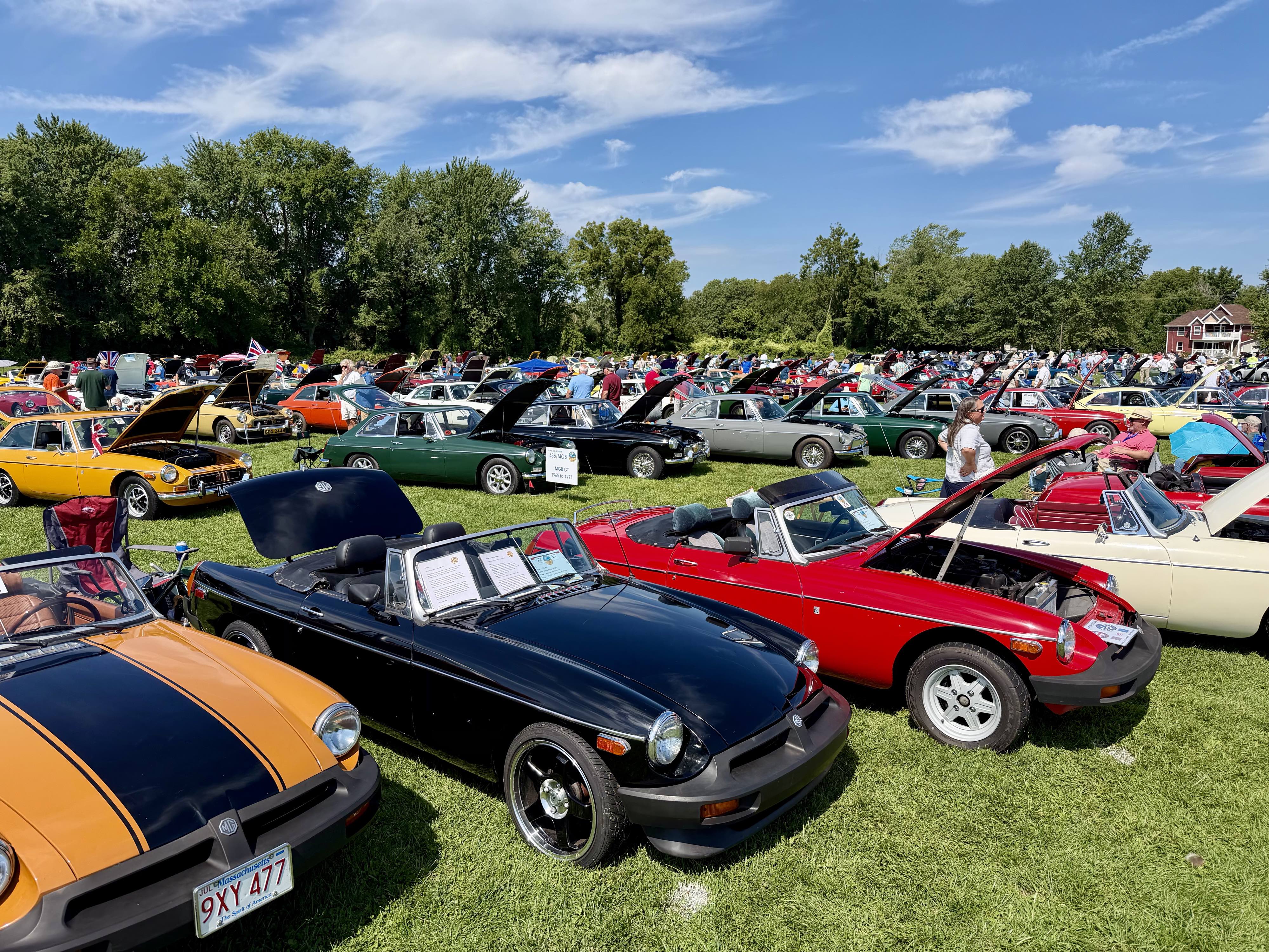 Several colorful rubber bumper MGB roadsters — including the black 1976 MGB nicknamed the FUN MG — in front of dozens hundreds of other colorful MGs on the show field at MG International 2026.￼