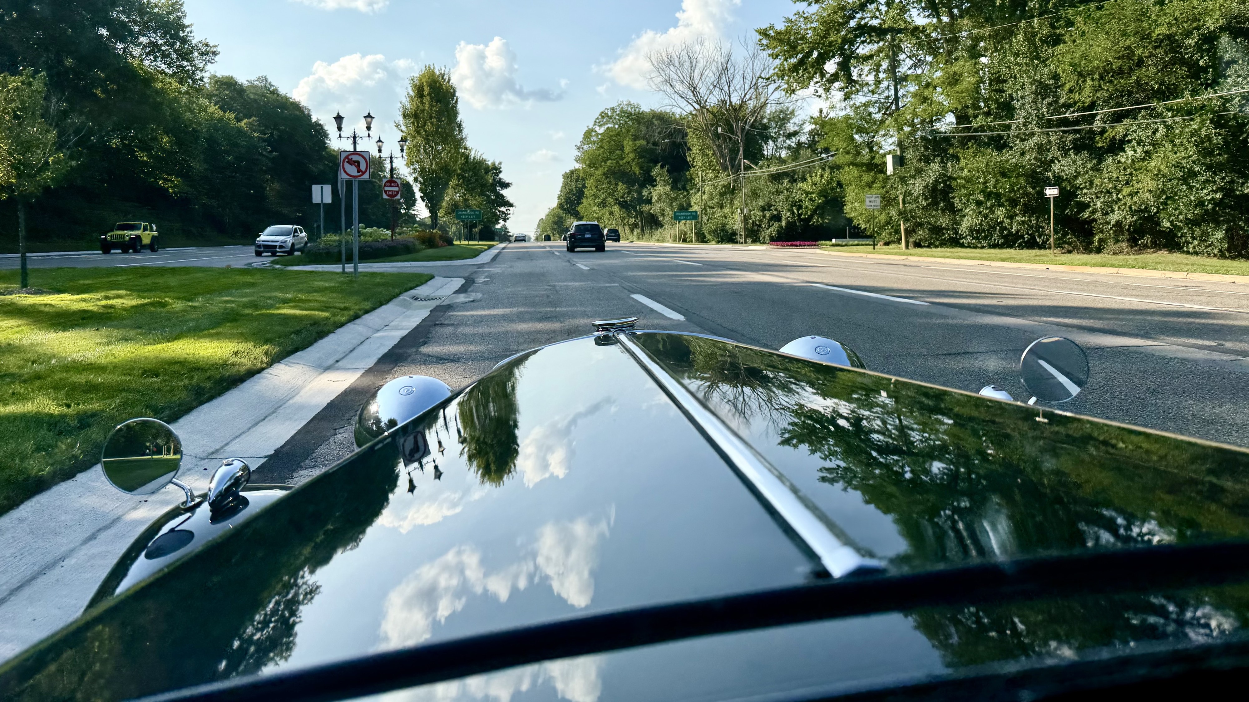 Photograph of the view down the hood of a dark green 1950 MGTD as it drives on a wooded boulevard in the summertime.
