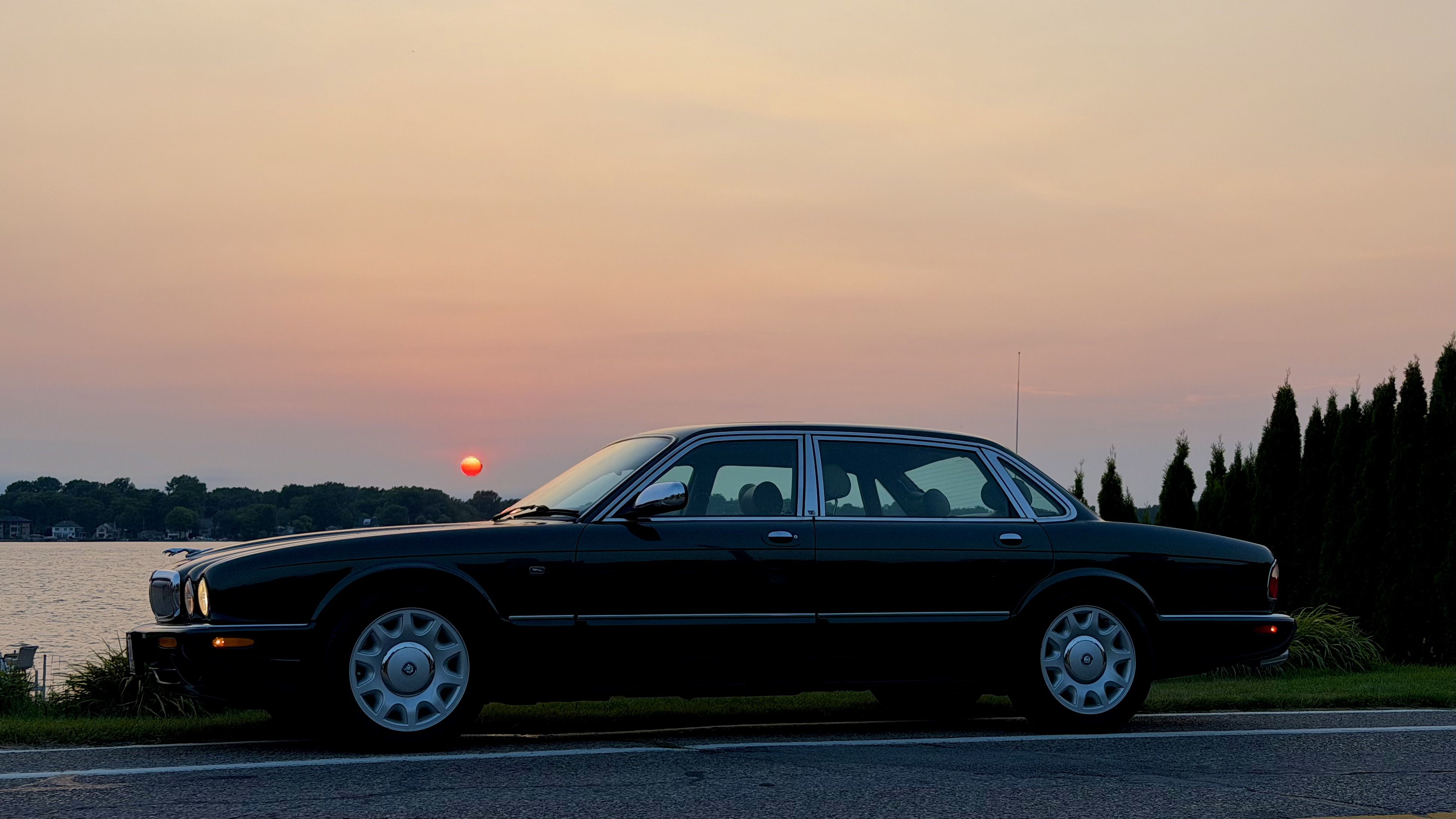 Photograph of a 1999 Jaguar Vanden Plas Supercharged sedan seen from the side with the front to the left. Behind it is a smoky sunset over a lake with a small bright red sun over the base of the windshield.