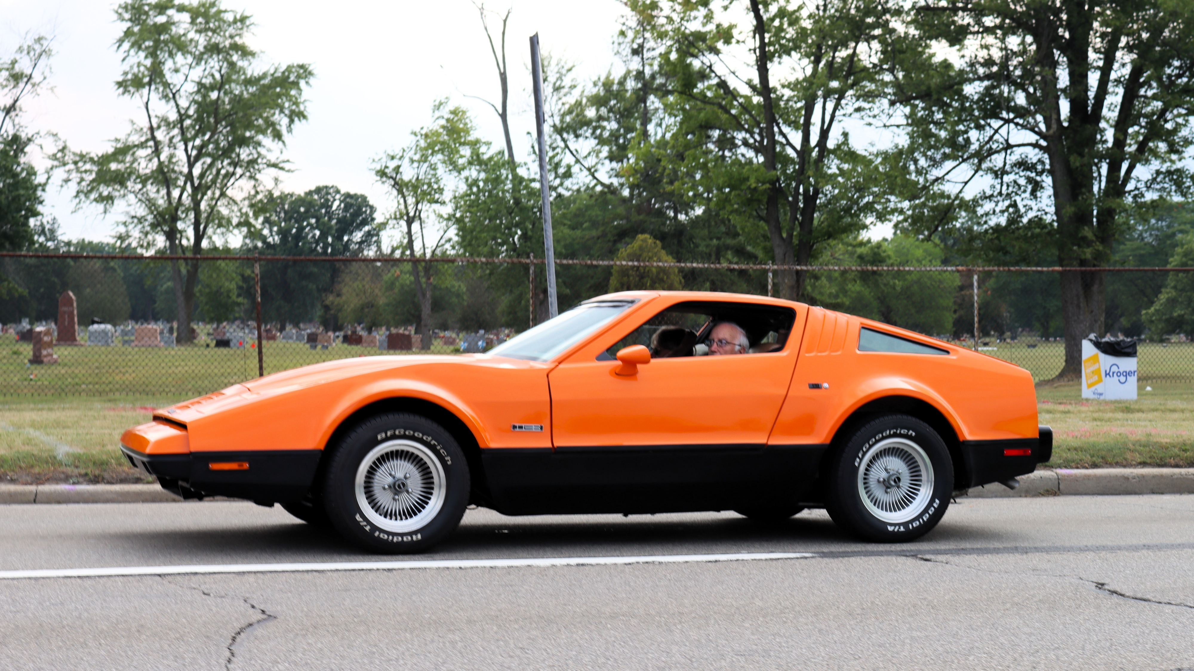 Photograph of an orange Bricklin sports car driving from right to left with a cemetery in the background.
