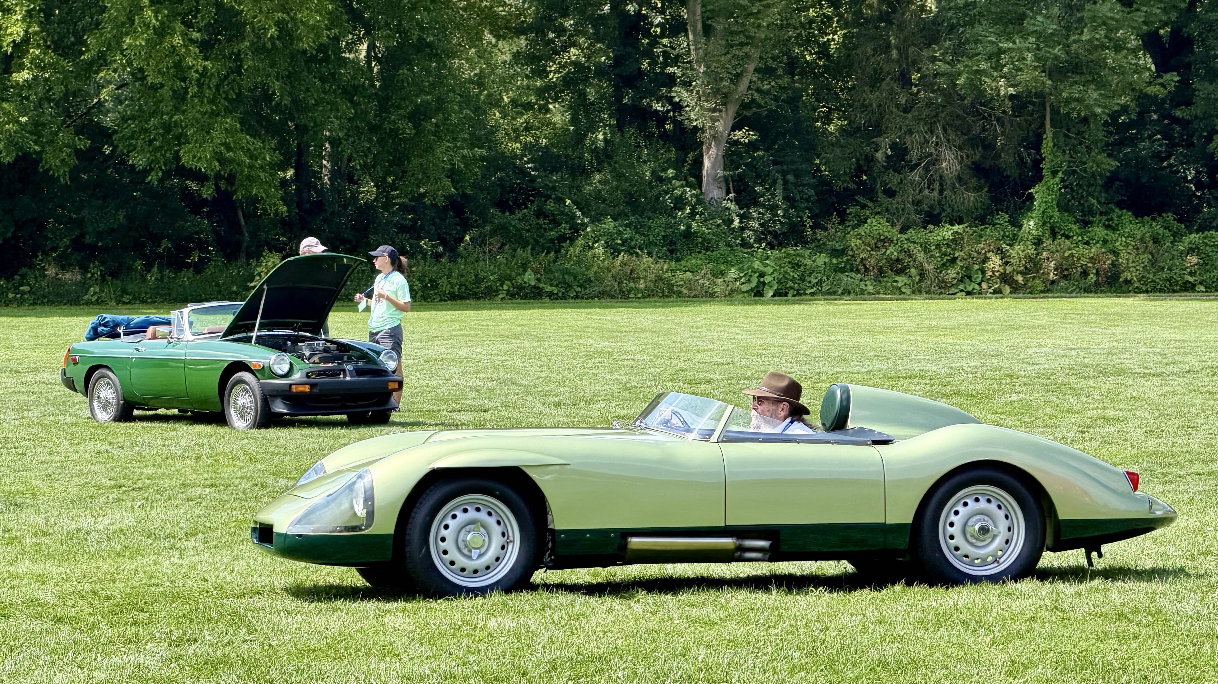 Photograph of a low-slung light green open-top race car driving across a grass field with a green MGB roadster in the background.