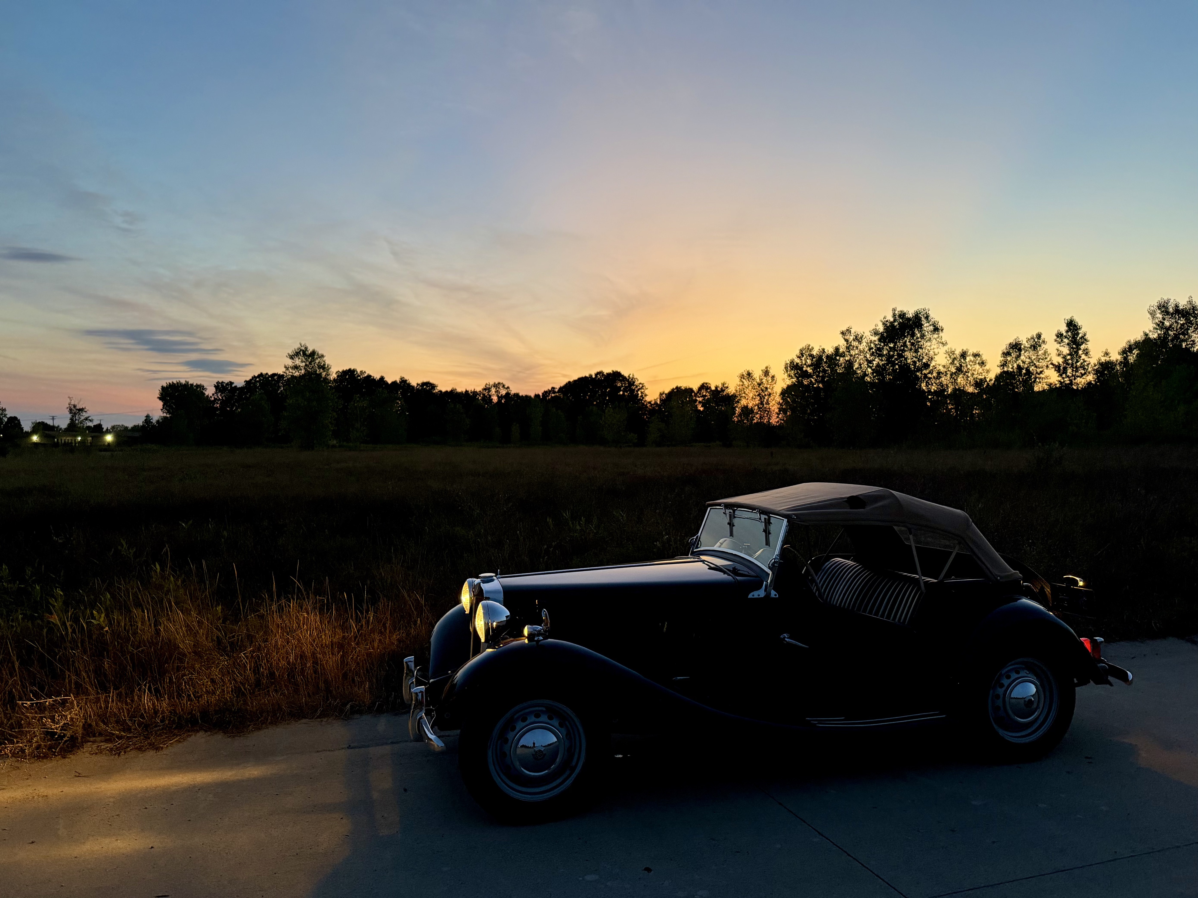 Photograph of an MG TD in front of a sunset with a field and tree line and silhouette behind it.