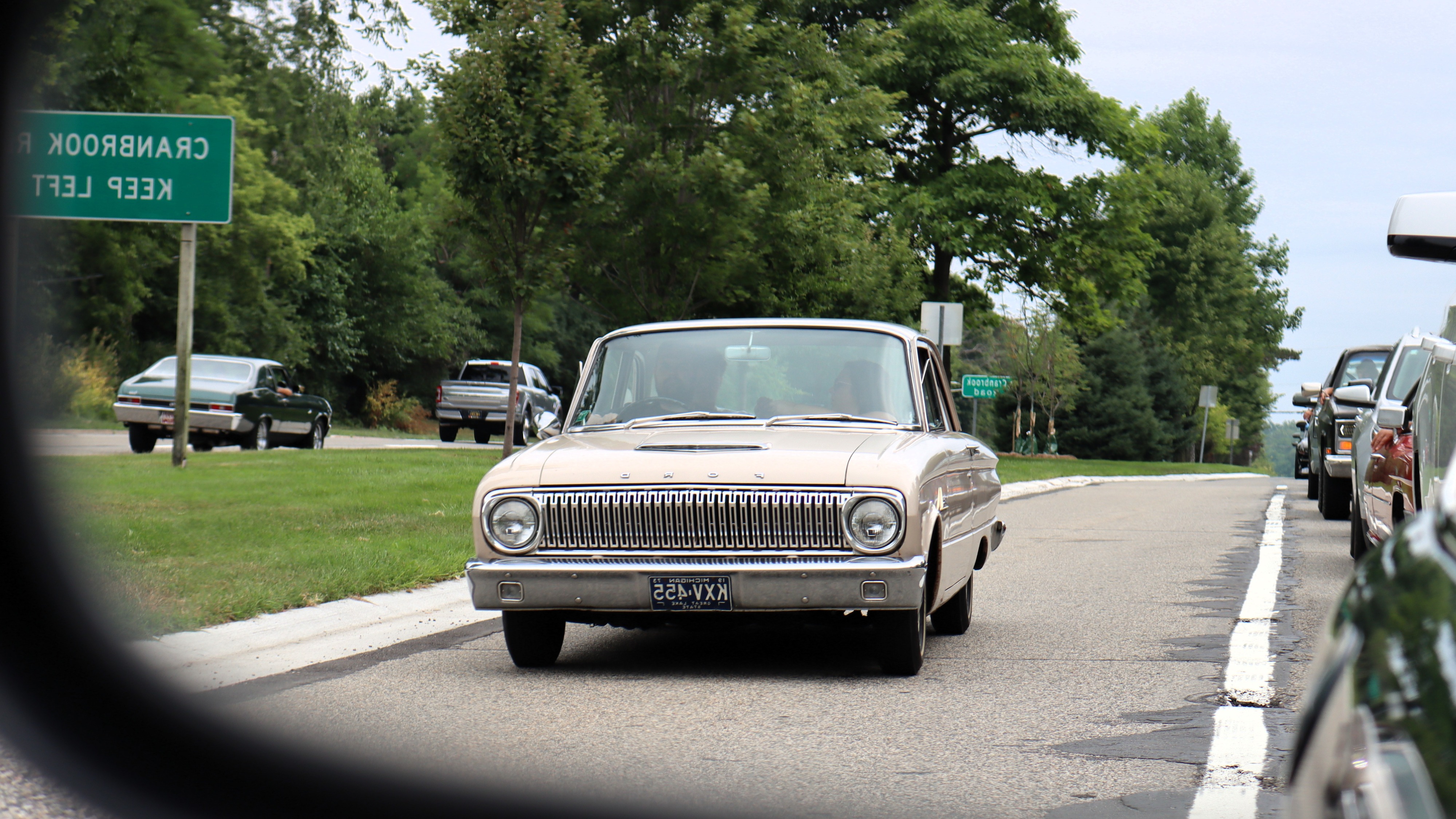 Photograph of an early 1960s Ford Falcon coming from behind in a clear traffic lane as seen in the left side mirror during the 2026 Dream Cruise.