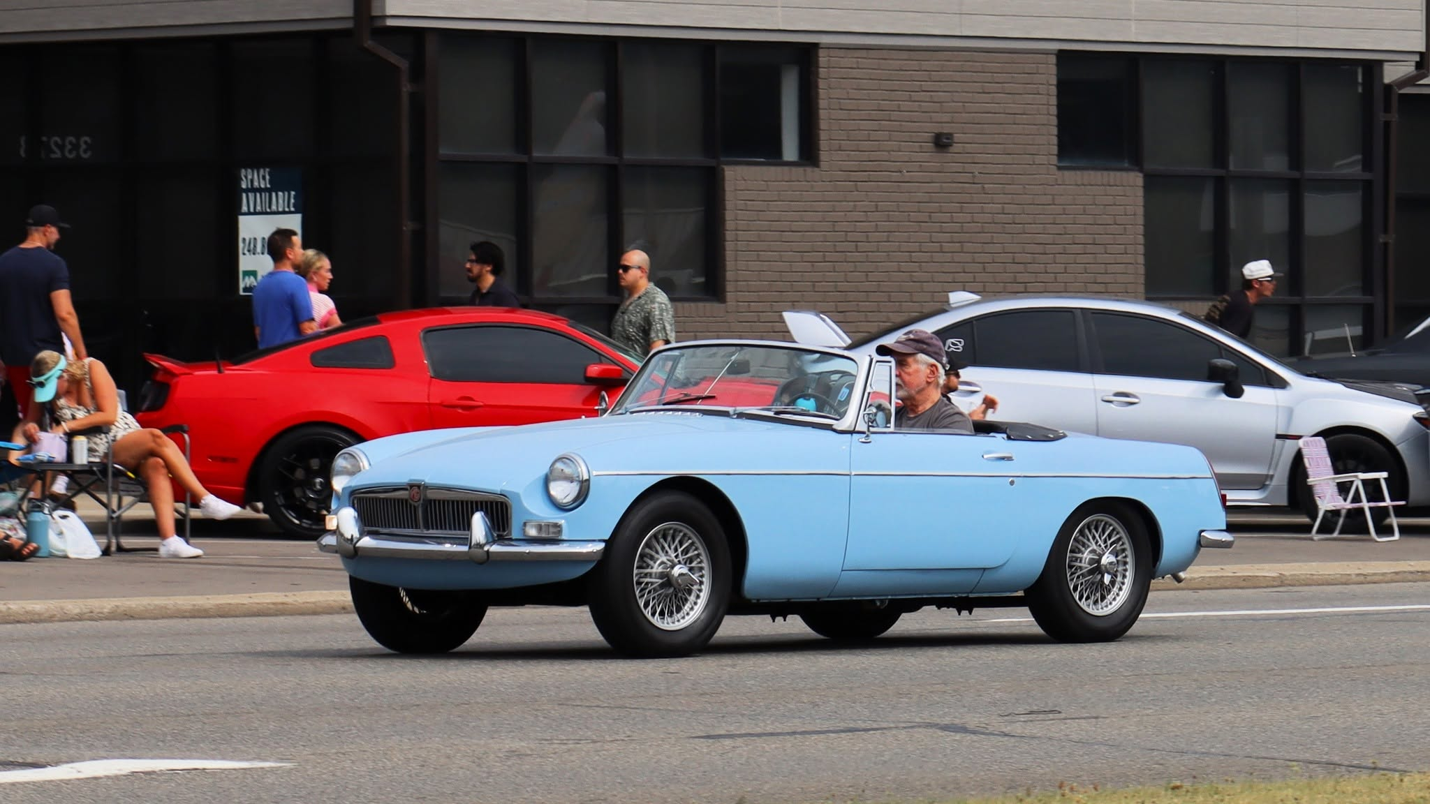 Photograph of a 1963-65 "pull handle" MGB in light blue (probably Iris Blue) driving down a street as seen from the front left.