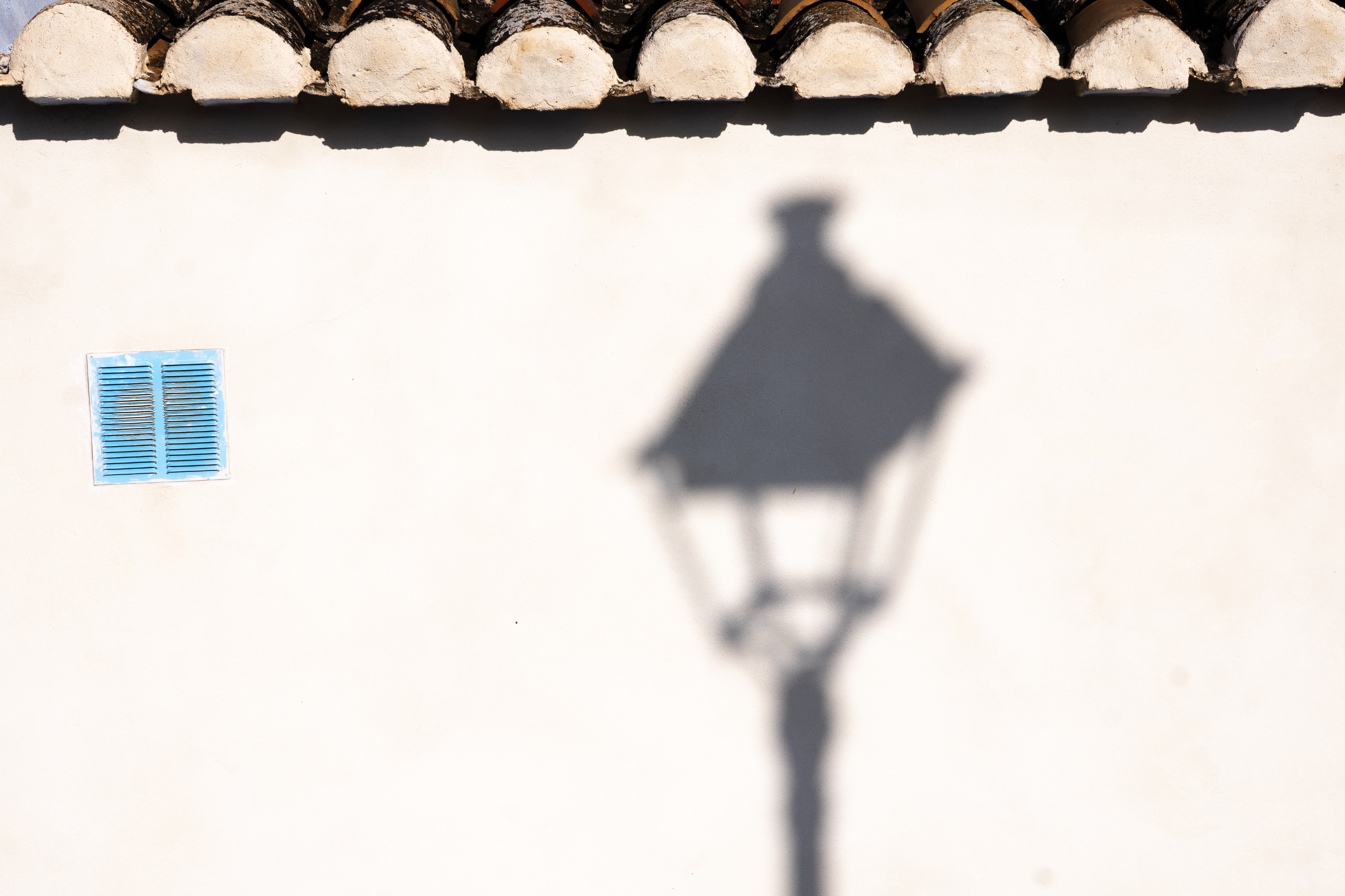 The photograph shows the shadow of a lamppost clearly outlined by the bright Spanish sunshine on a stark, white wall.  To the left on the wall is a blue ventilation grill.  The edges of the Spanish roof tiles can be seen at the top edge of the photo.  